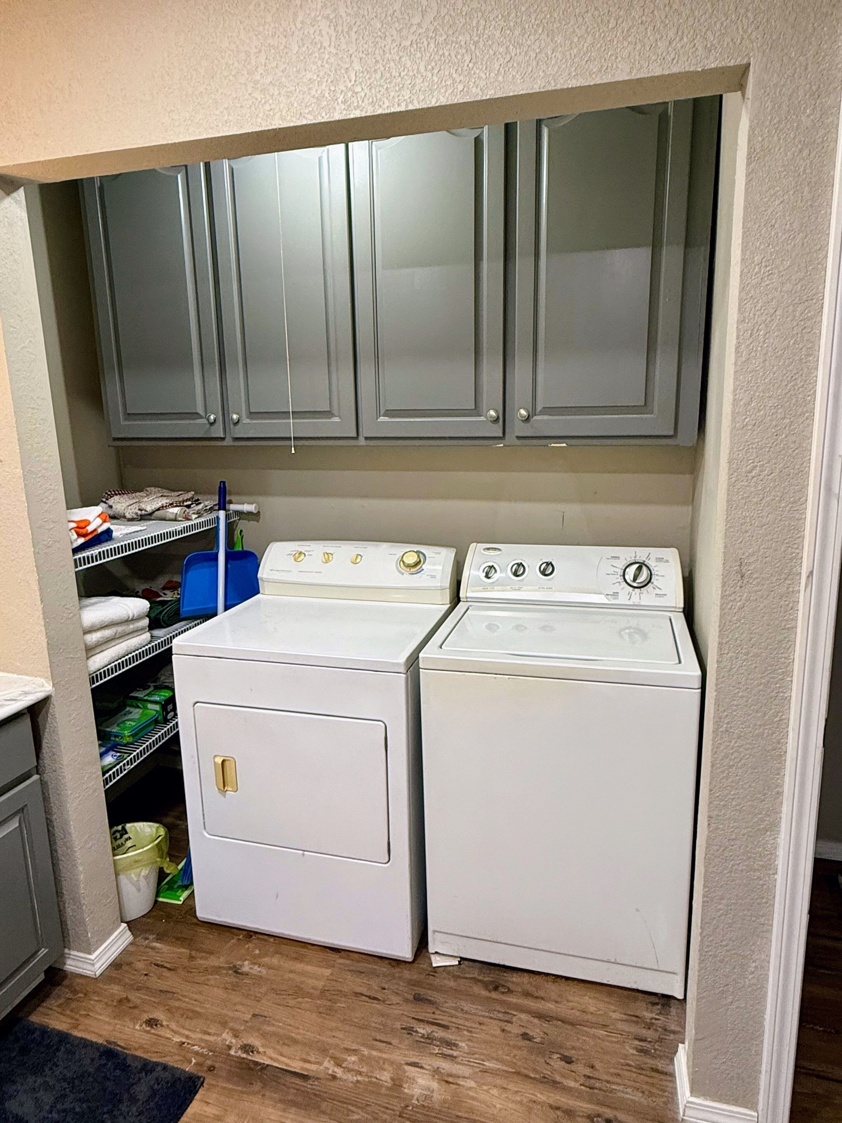 A compact laundry room with two white front-loading washers and dryers, grey cabinets above, and a shelf with folded towels and cleaning supplies.