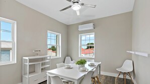 A dining area with a white table, chairs, and a ceiling fan. There are windows with views of buildings outside. - Queen's Promise - King Suite - AC - Walkable (Christiansted)