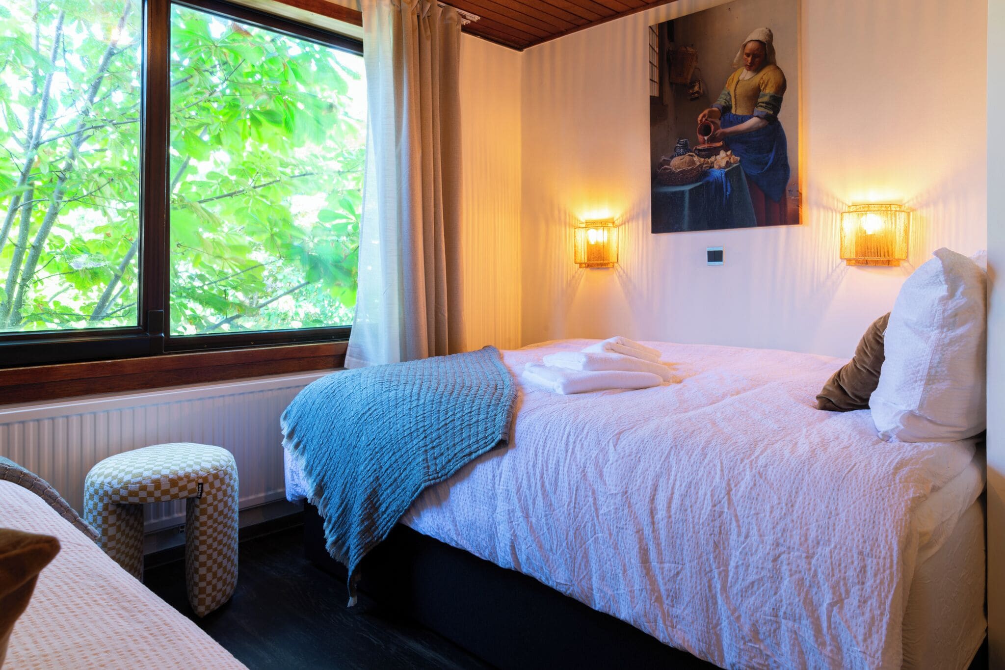 A bedroom with a bed, a window with curtains, a wall-mounted light, and a painting.