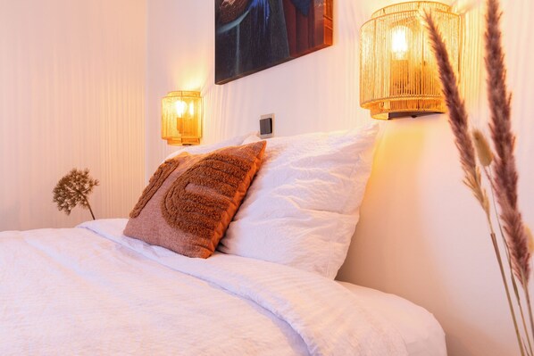 Una cama bien hecha con sábanas blancas y un cojín marrón, dos lámparas de pared y un cuadro sobre la cama.