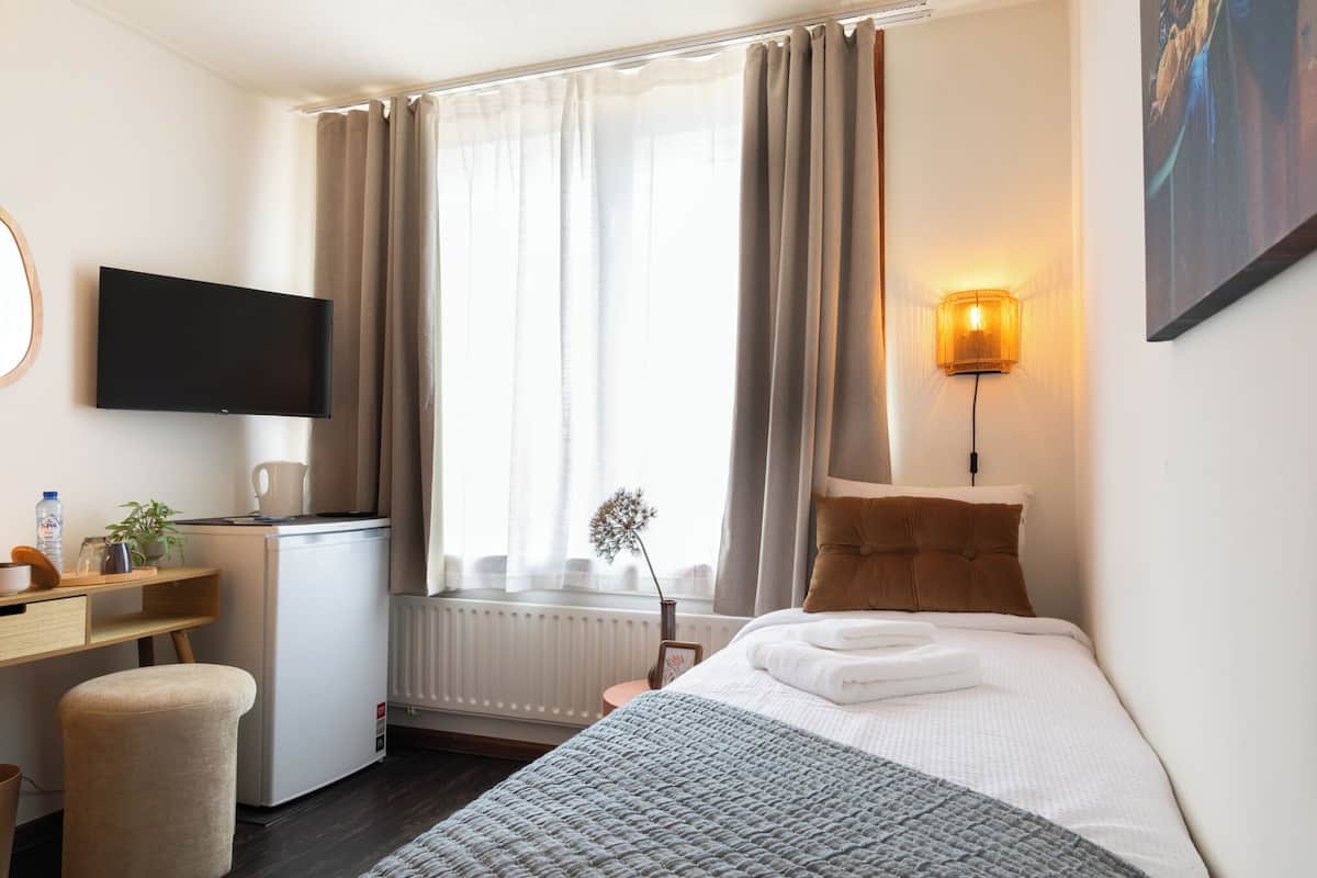 Une chambre d’hôtel comprenant un lit, un bureau avec un ordinateur, une télévision, une fenêtre avec des rideaux, une lampe fixée au mur et un radiateur.