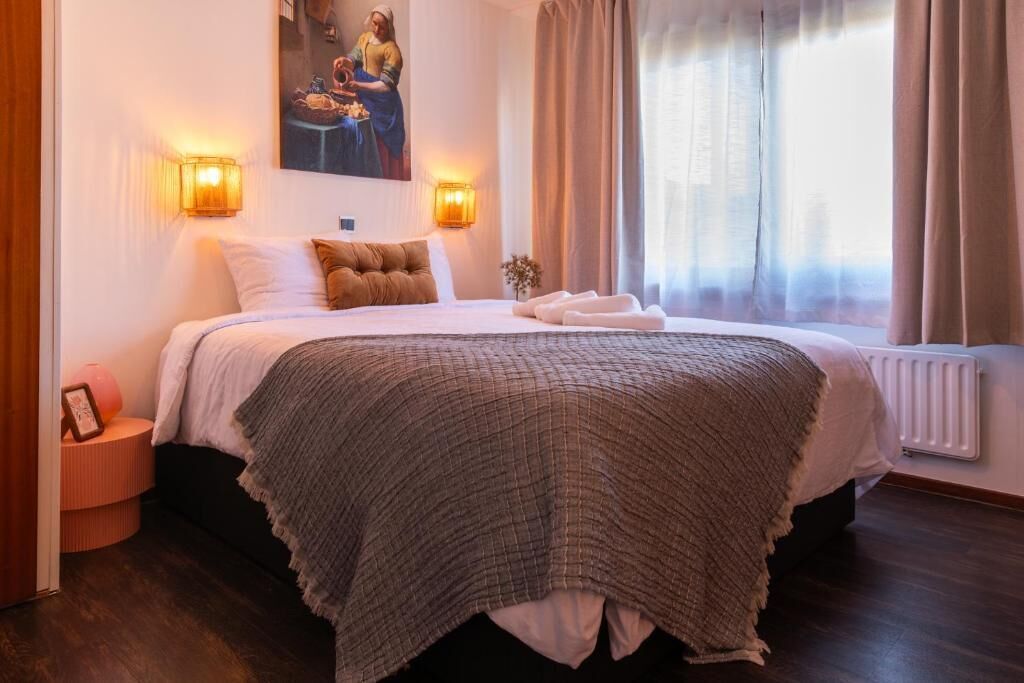 Una cama bien hecha con un edredón texturizado, dos lámparas de noche, un cuadro enmarcado en la pared y una ventana con cortinas.