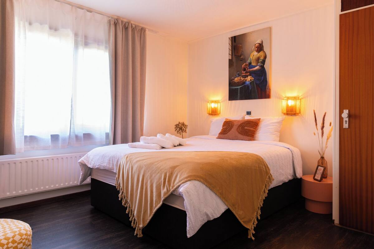 Une chambre d’hôtel avec un grand lit, une fenêtre avec des rideaux, un tableau au mur et deux lampes fixées au mur.