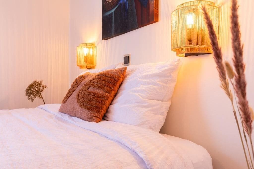 A neatly made bed with white linens and a brown throw pillow, flanked by two wall-mounted lamps with a warm glow.