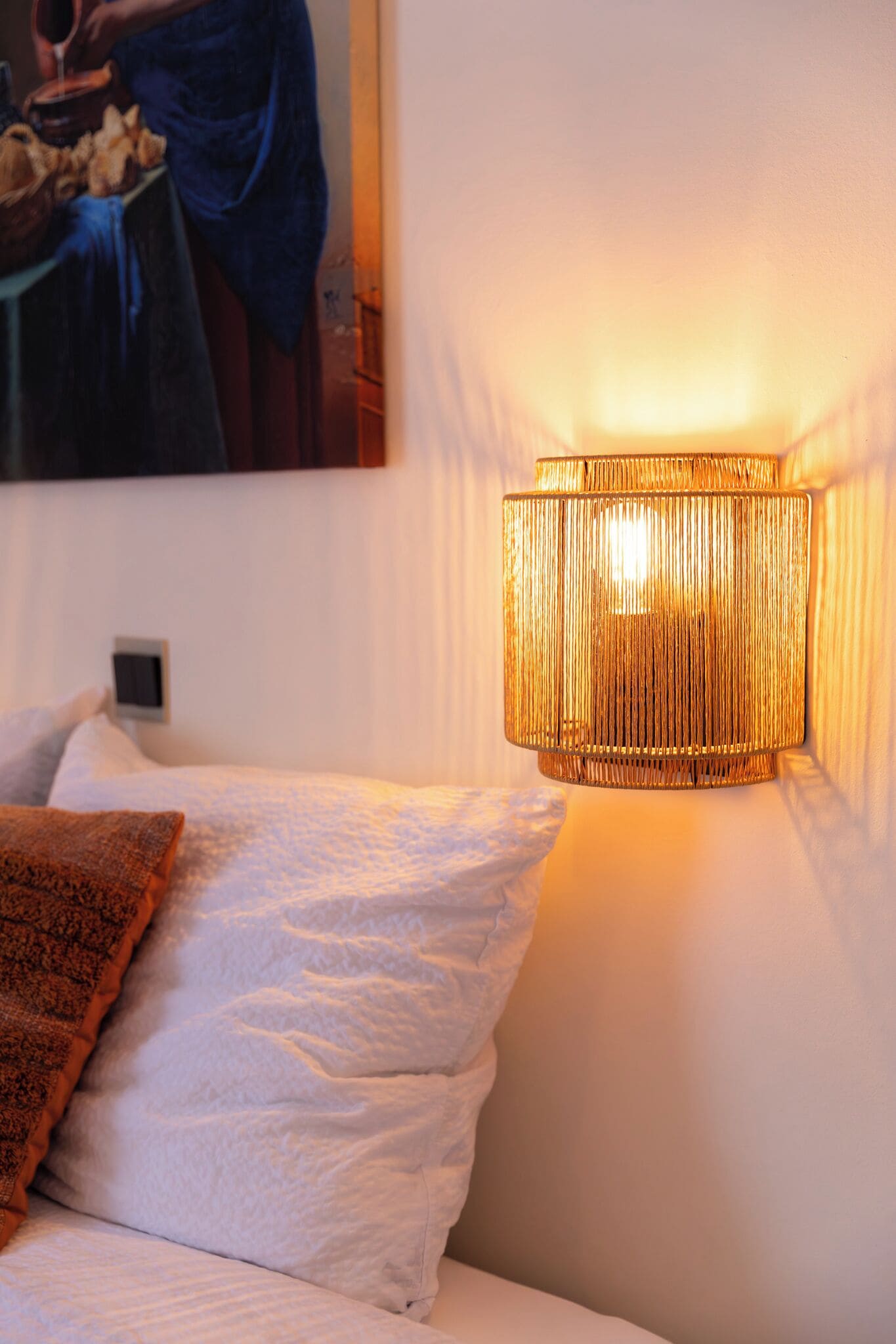 Una cama con sábanas blancas y un plaid marrón, una lámpara de pared y un cuadro sobre la cama.
