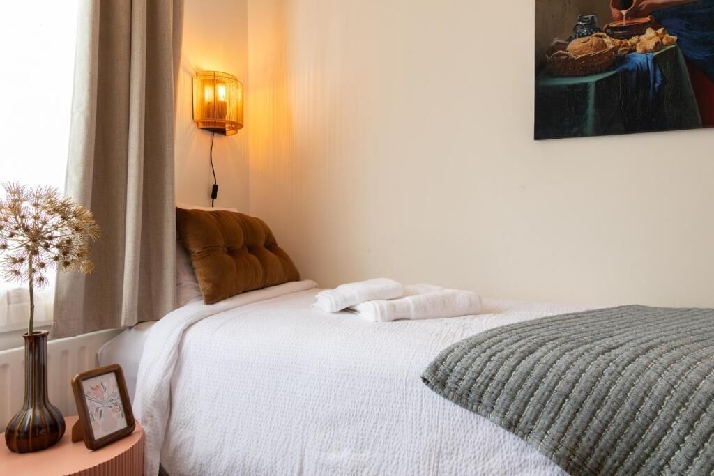 A neatly made bed with white linens, a gray throw blanket, and a brown pillow. There is a vase with dried flowers on a bedside table, and a framed picture beside it. A wall-mounted lamp provides soft lighting.