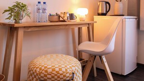 A compact hotel room with a wooden desk, a white chair, a checkered stool, a television mounted on the wall, a small table with a lamp, a plant, and a refrigerator.