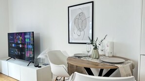 A modern living room with a flat-screen TV on a white cabinet, a round wooden dining table with white chairs, and a framed abstract artwork on the wall.