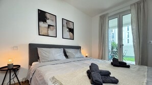 A modern hotel room with a large bed, two abstract paintings on the wall, a bedside table with a lamp, and a view of the outside through a sliding glass door.