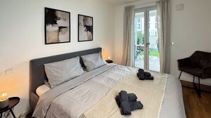 A modern bedroom with a large bed, two abstract paintings, a wicker ceiling light, a view of the outdoors through a sliding door, and a dark armchair.
