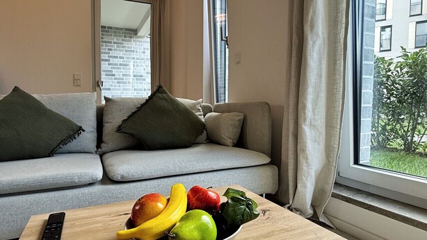 A living room with a grey sofa, a wooden table with a bowl of fruit, and a window with curtains.