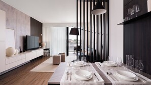 Un espace de repas moderne avec une longue table dressée pour un repas, un téléviseur sur le côté gauche et une vue sur l’extérieur grâce à une grande fenêtre.
