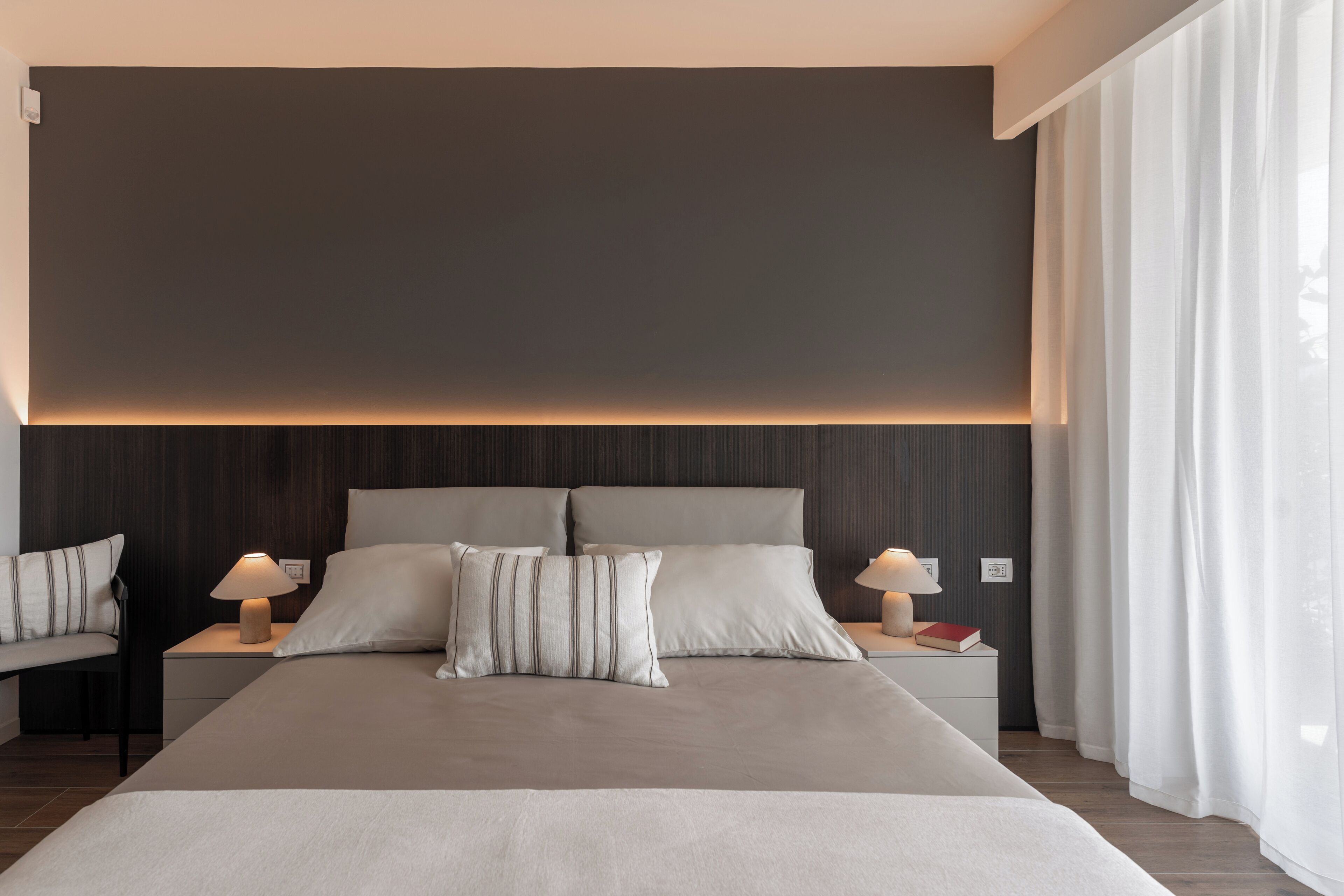 Una habitación de hotel moderna con una cama grande, dos mesitas de noche con lámparas, y vistas al exterior a través de cortinas translúcidas.