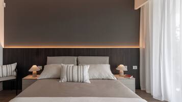 Una habitación de hotel moderna con una cama grande, dos mesitas de noche con lámparas, y vistas al exterior a través de cortinas translúcidas.