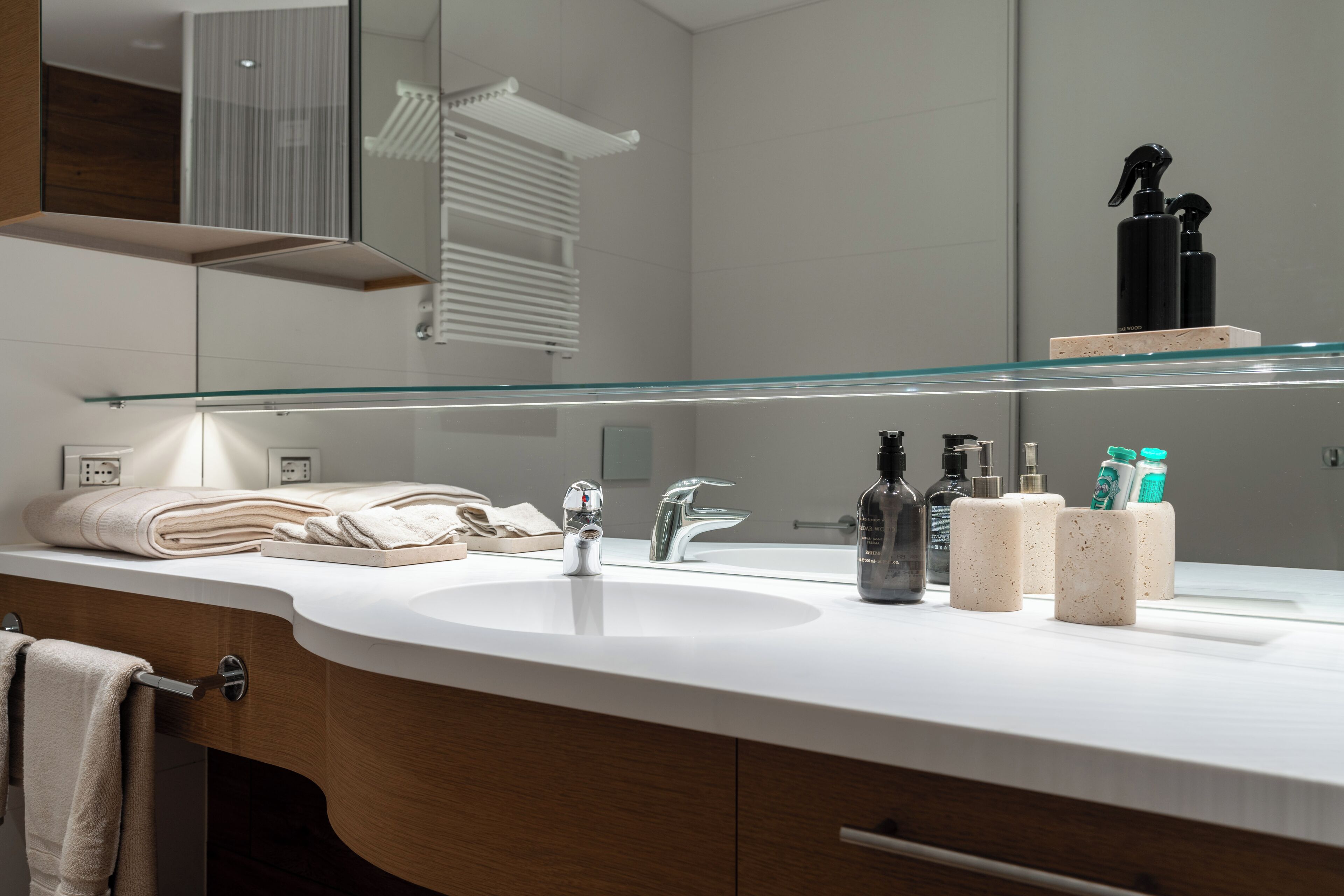 Un baño moderno con un lavabo blanco, dos grifos, un estante de vidrio sobre el lavabo y un perchero con toallas dobladas.