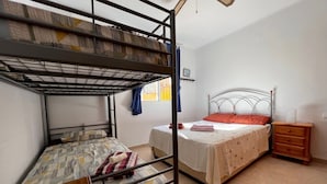 Egy kollégiumi szoba emeletes ágyakkal, egy egyszerű ággyal, egy éjjeliszekrénnyel és egy mennyezeti ventilátorral.