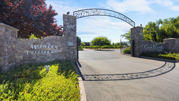 A winery tasting room entrance with stone walls, a metal archway, and a paved path leading inside.