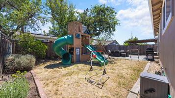 Ein Garten mit einem hölzernen Spielhäuschen, einer grünen Rutsche und einem umzäunten Bereich.