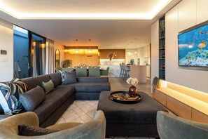 A modern living room with a sectional sofa, a coffee table with decorative items, a TV mounted on the wall, and a kitchen area in the background.