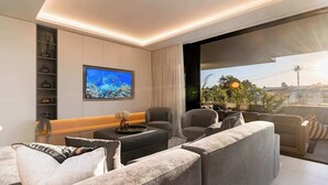 A modern living room with a large flat-screen TV, grey sofas, a coffee table, and a view of the city through a large window. - Walk to beach, Pool, Camps Bay -Terrace Royale Apt (Cape Town)