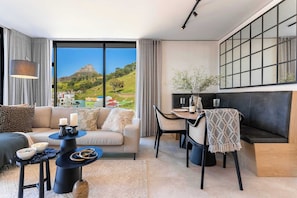 A modern living room with a sofa, dining table, and chairs, large windows with a view of a hill and buildings, and a black-framed artwork on the wall. - Modern, Gorgeous Views, Bo-Kaap - Vista Luxe Apt (Cape Town)