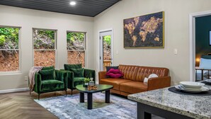 A modern living room with a green velvet sofa, a brown leather couch, and a coffee table. There are large windows with a view of greenery outside.