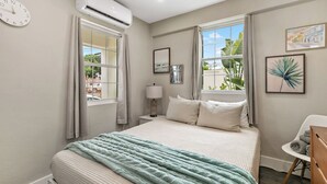 A bedroom with a bed, an air conditioner, a window with curtains, a bedside lamp, and artwork on the wall.