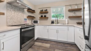 A modern kitchen with white cabinets, a stainless steel range hood, a microwave, and wooden shelves.