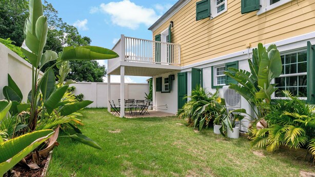 A yellow building with a balcony and green shutters, surrounded by lush greenery and a well-maintained lawn.