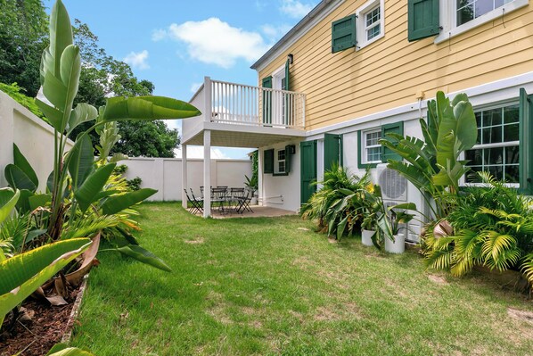 A yellow building with a balcony and green shutters, surrounded by lush greenery and a well-maintained lawn.
