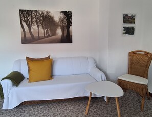 A room with a white sofa, a mustard-colored pillow, a small white table, a wicker chair, and a framed picture on the wall.
