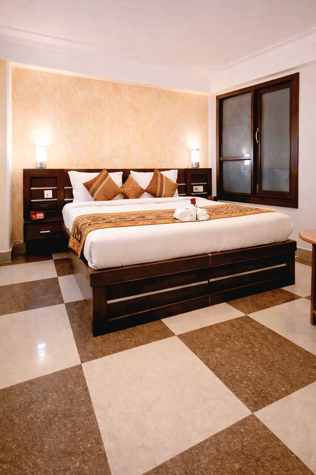 A hotel room with a checkered floor, a bed with a wooden headboard, two bedside tables, and a large window with a view.