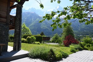 A wooden cabin with stone pillars, a garden with various plants, and a view of snow-capped mountains.