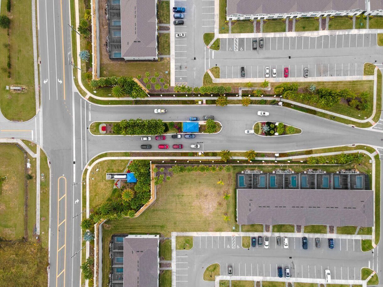 Une vue aérienne d’un parking comportant plusieurs rangées de voitures, un espace paysager central et des bâtiments en arrière-plan.