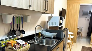 A modern kitchen with wooden cabinets, a black countertop, and a built-in sink. There are various kitchen utensils hanging on the wall, a microwave, and a small refrigerator. - Crizzy's Hub Sariel @ Shore Residences (Pasay City, Metro Manila)