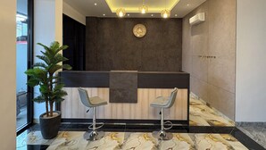 Uma área de recepção com piso de mármore, balcão de recepção, dois banquinhos de bar, uma planta em vaso e um relógio de parede.