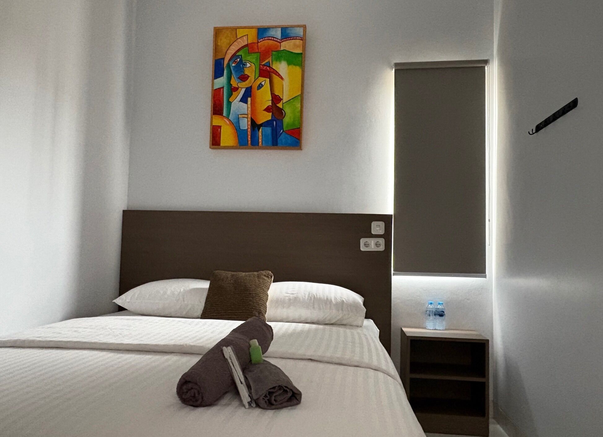 A hotel room with a bed, pillows, a towel, a painting, a nightstand, and a door.