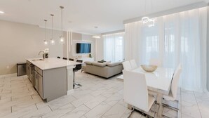 Una cocina moderna con una isla central, un comedor con mesa y sillas blancas, y una sala de estar con sofá y televisor.