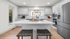 A modern kitchen with white cabinetry, a central island, two bar stools, and stainless steel appliances. - Heated Pool! MiniGolf! Hot Tub! Fire Pit! Lakefront! (Hot Springs)