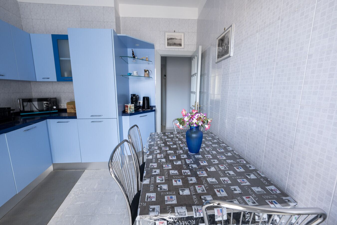 Una cocina compacta con armarios azules, una mesa de comedor con mantel estampado de periódico y un jarrón con flores.