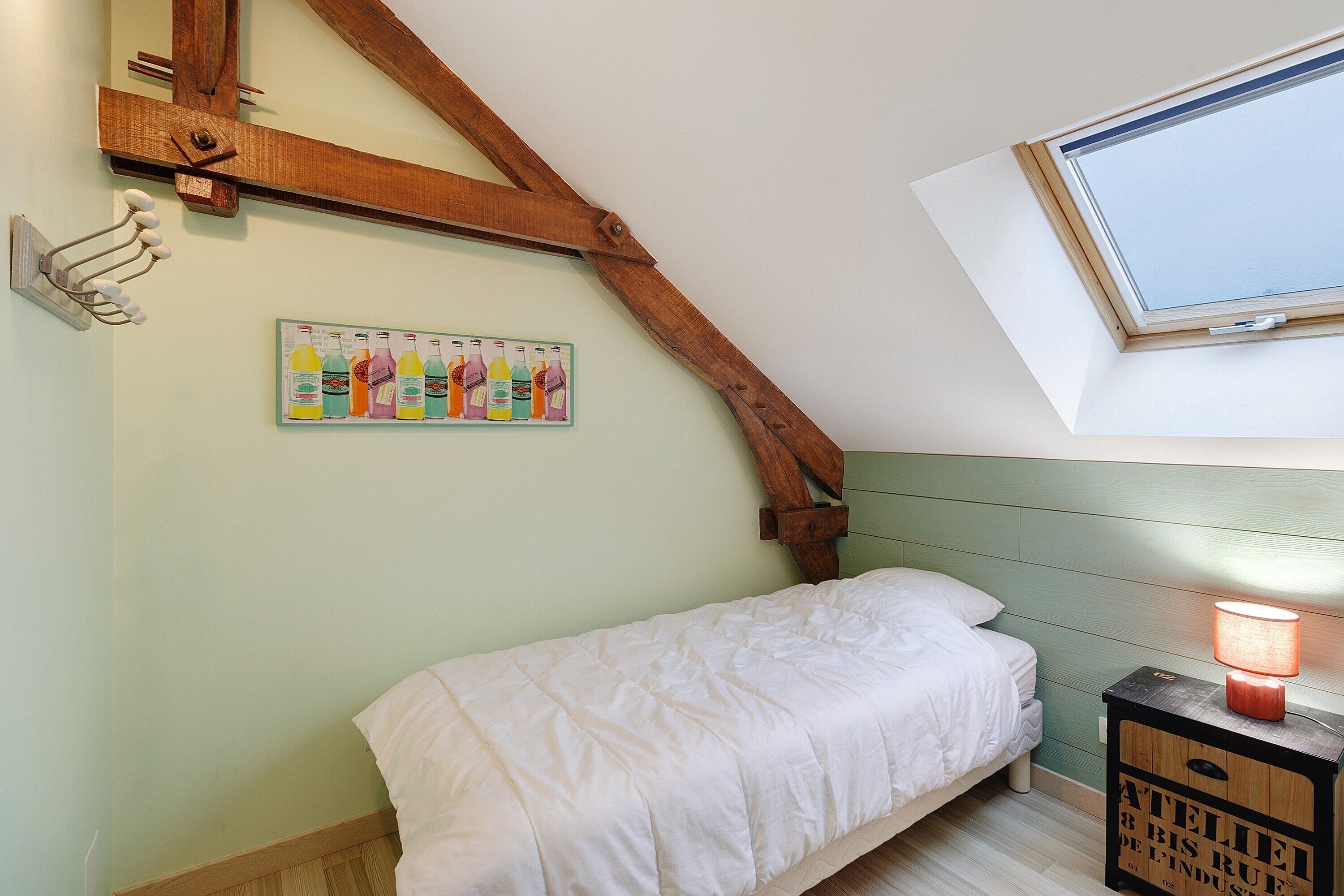 A bedroom with a bed, a nightstand with a lamp, a wooden beam ceiling, a window, and a wall-mounted artwork.
