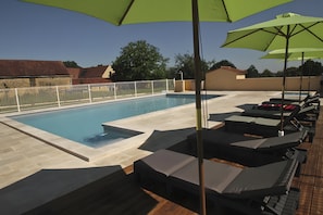 A swimming pool with lounge chairs and umbrellas, surrounded by a fence and a building with a tiled roof. - Holiday Home with Shared Pool, Wi-Fi and Air Conditioning (Saint-Geniès)