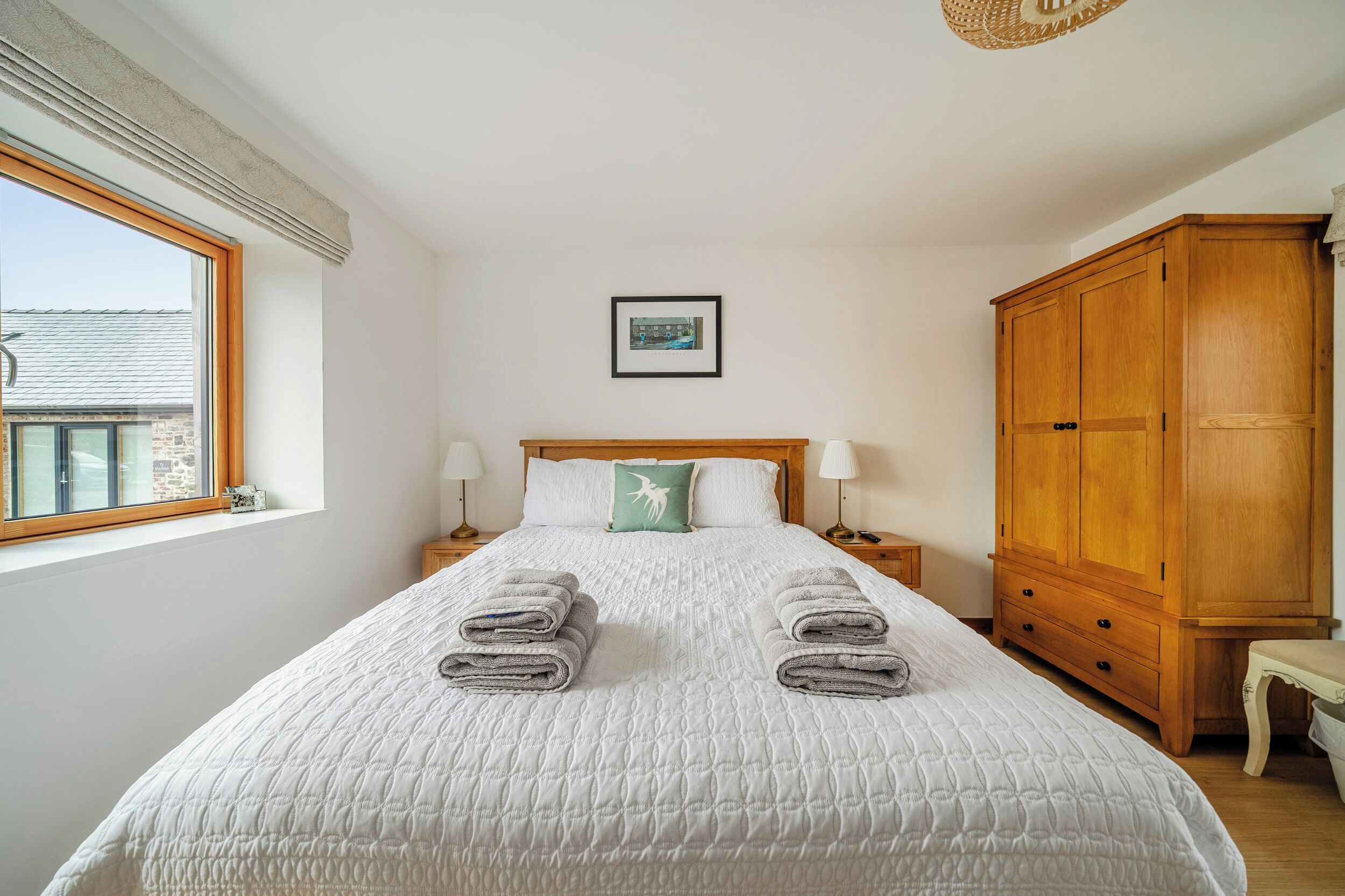 Un dormitorio con una cama, un armario de madera, un cuadro en la pared y una ventana con vistas a edificios.