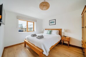 A bedroom with a wooden bed, a bedside table with a lamp, a window with a view of buildings, and a framed picture on the wall. - Hen Penmynydd (Holyhead)