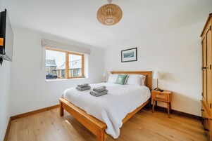 Un dormitorio con cama de madera, una mesita de noche con lámpara, una ventana con vistas a edificios y un cuadro enmarcado en la pared.