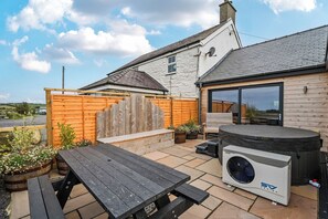 A patio area with a wooden table, outdoor seating, a hot tub, and a large fan. - Hen Penmynydd (Holyhead)