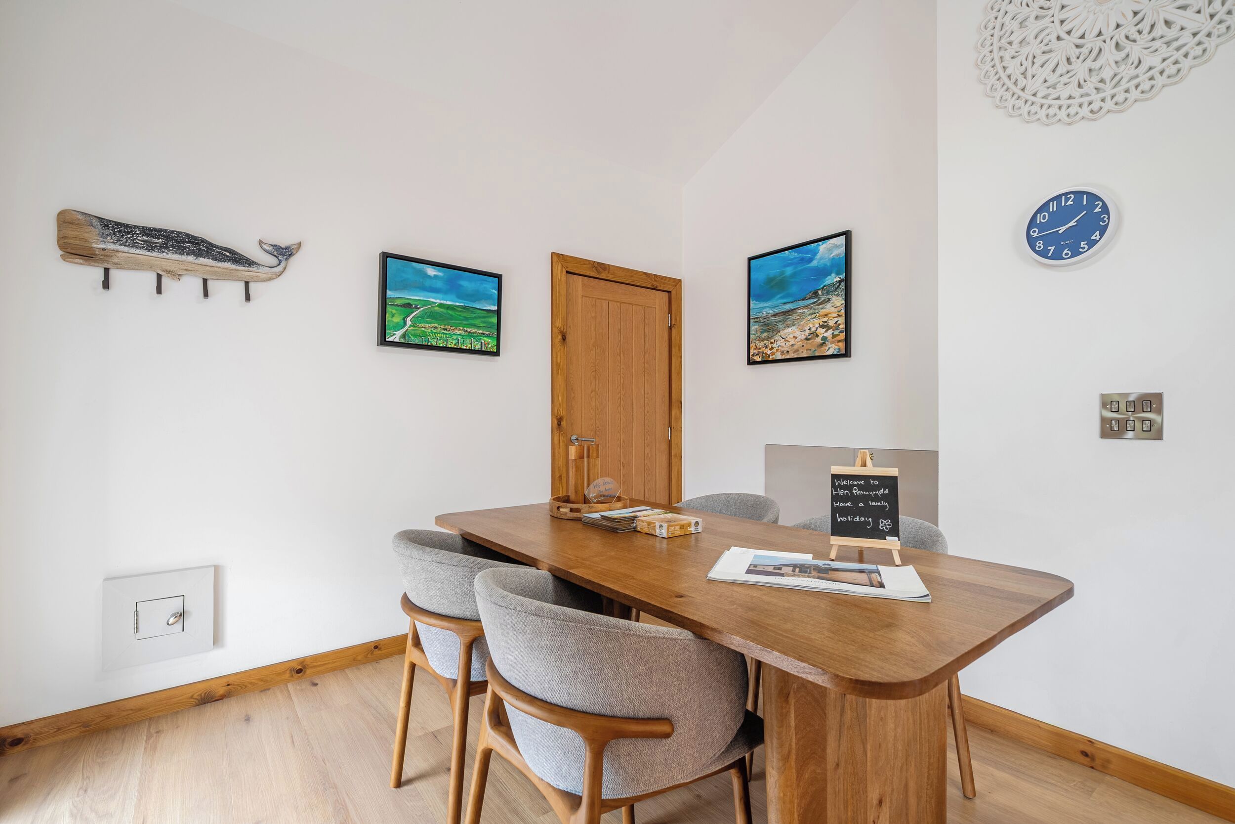 A dining area with a wooden table, four grey chairs, two framed landscape paintings, a wooden door, and a wall clock.