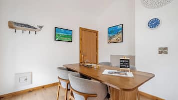 A dining area with a wooden table, four grey chairs, two framed landscape paintings, a wooden door, and a wall clock.