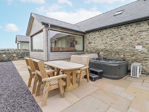 A wooden outdoor seating area with a table and chairs, a hot tub, and a stone wall. - 2 Trem Y Mor (Holyhead)