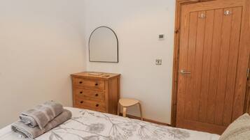 Une chambre à coucher comprenant un lit, une commode en bois, un petit tabouret, une porte en bois et un miroir fixé au mur.